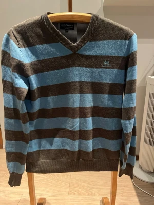 Randig grå och blå tröja Jean Paul - Säljer en grå och ljusblå randig tröja från Jean Paul i 100% bomull. Tröjan har v-ringning, långa ärmar och ribbade muddar. Perfekt för dig som gillar klassisk stil med en twist. Märkeslogga broderad på bröstet.