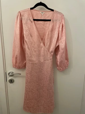 Rosa omlottklänning från Object - Söt rosa omlottklänning från Object med subtilt blommönster i tyget. Klänningen har v-ringning, trekvartsärmar med resår och knytband i midjan. Materialet har en lätt glansig finish som ger en lyxig känsla.