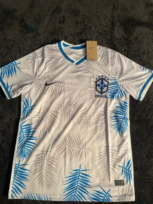 Vit och blå Brasilien Nike fotbollströja - Snygg Brasilien fotbollströja från Nike i vitt med blå detaljer och palmbladsmönster. Tröjan har V-ringad krage med blå kant, CBF-logga på bröstet och Dri-FIT-material som andas. Perfekt för dig som älskar fotboll och vill sticka ut på planen.