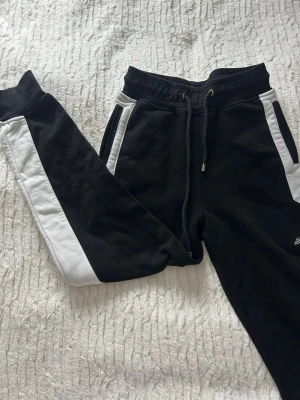 Svarta mjukisbyxor från Elly Pistol - Svarta mjukisbyxor från Elly Pistol med vita paneler längs sidorna och broderad logga på benet. Byxorna har snörning i midjan, ribbade muddar vid benslut och två fickor. Tillverkade i mjukt bomullsmaterial för extra komfort. Finns arr köpa matchande Hoddie till så ett sett