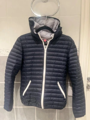 Mörkblå Colmar pufferjacka 14Y  - Snygg mörkblå pufferjacka från Colmar med vit dragkedja och två fickor med dragkedja. Jackan har huva och quiltad design som ger en cool look. Perfekt för dig som vill ha en lätt och varm jacka med stil. Colmar-logga på ärmen.