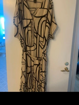 Beige och svart mönstrad klänning - Säljer en beige klänning från Zara med svart abstrakt mönster. Klänningen har korta ärmar, v-ringning och ett bälte i midjan för snygg passform. Materialet är mjukt och lätt, perfekt för varmare dagar. Passar dig som gillar unik design och bekväma plagg.