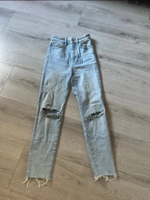 Ljusblå mom jeans från Stradivarius - Säljer ett par ljusblå mom jeans från Stradivarius i storlek 32. De har hög midja, raka ben och slitna detaljer med hål på båda knäna samt fransiga benslut. 