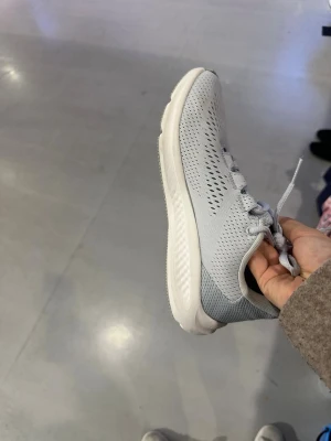 Ljusgrå sneakers från Under Armour - Snygga ljusgrå sneakers från Under Armour med ventilerande meshmaterial och vit platt sula. Skorna har snörning och ett stort Under Armour-logotyp på sidan. Perfekta för träning eller chill dagar. Modern design med rund tå och bekväm passform.