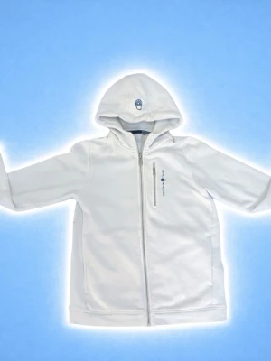 Vit vindjacka med huva från Sailracing - Snygg vit vindjacka från Sail racing  med huva och hel dragkedja framtill. Jackan har en bröstficka med dragkedja och diskret logga på bröstet. Perfekt för blåsiga dagar och enkel att matcha med det mesta. Det är 170 för barn men jag har tvättat den vissa gånger så den är ca 158/164