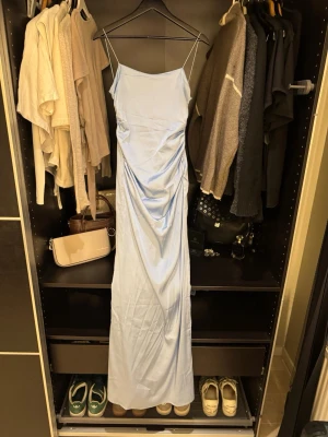 Ljusblå balklänning - Supersnygg ljusblå långklänning med tunna axelband och draperad detalj på sidan. Klänningen har en slät, glansig yta som ger en lyxig känsla och en öppen rygg. Perfekt för dig som gillar stilrena och eleganta plagg med en modern twist. Den har även en slit.