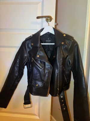 Svart skinnjacka från Stradivarius XS - Snygg svart skinnjacka från Stradivarius i storlek XS. Jackan har flera silvriga dragkedjor, knappar i metall och ett bälte i midjan. Klassisk bikerstil med lång ärm och coola detaljer som fickor och axelklaffar. Perfekt för att lyfta din outfit.