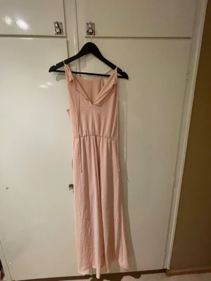 Ljusrosa långklänning  - Superfin ljusrosa långklänning med rak framsida och smala axelband. Klänningen har markerad midja och är tillverkad i ett mjukt, lätt tyg som faller snyggt. Perfekt för sommarens alla tillfällen!