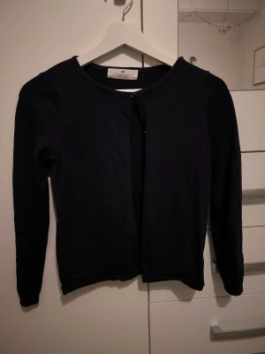 Svart kofta från H&M - En enkel svart kofta från H&M med rund halsringning och lång ärm. Koftan har en knapp högst upp och är tillverkad i mjukt material som känns skönt mot huden. Perfekt att slänga över en topp när det blir lite kyligt.
