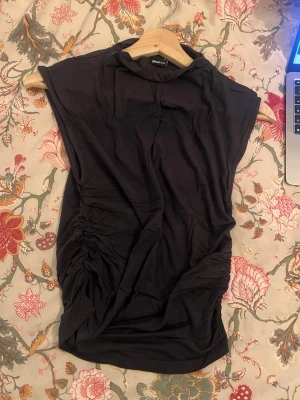 Svart ärmlös topp från Gina Tricot - Säljer en svart ärmlös topp från Gina Tricot med hög hals och snygga rynkade detaljer på sidorna. Toppen är i mjukt material och har en clean, modern look som passar till många olika outfits.