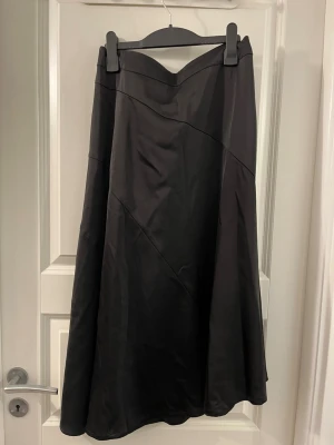 Svart långkjol från Vero Moda - Säljer en svart långkjol från Vero Moda i storlek M. Kjolen har en snygg, svepande A-linjeform och bred resår i midjan för skön passform. Materialet är mjukt och har en lätt glansig yta med diskreta sömdetaljer som ger extra stil.