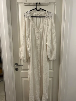 Vit långklänning med spets från H&M - Säljer en vit långklänning från H&M med v-ringning och broderade spetsdetaljer längs halsringning, axlar och ärmar. Klänningen har långa, luftiga ärmar och knappar framtill. Materialet är mjukt och lätt bomull, perfekt för varma dagar.