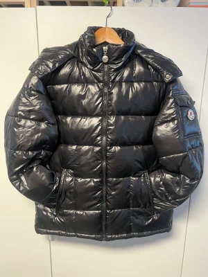 Svart pufferjacka från Feshion - Säljer en svart pufferjacka från Fashion med glansig finish och vadderad design. Jackan har huva, dragkedja framtill, fickor med dragkedja och en patch på ärmen. Perfekt för kalla dagar och riktigt snygg till streetwear-stilen. Storleken är XL men känns mer som en L då jackan är lite i storlek. Fashion loggan på vänster armen kan tas av och på. Frakt sker inom 24h! 💨📦