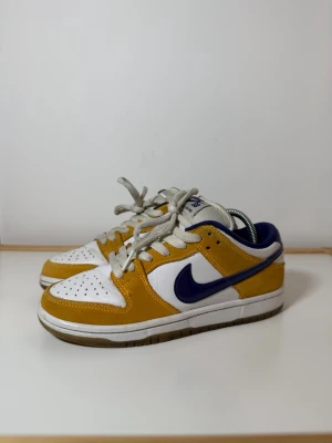 Nike SB Dunk Low laser orange sneaker  - Nike SB Dunk Low sneakers med vit läderbas, gula mockadetaljer och mörkblå swoosh. Skorna har klassisk snörning, perforerad tå och gråa skosnören. Perfekta för dig som gillar streetstyle och vill sticka ut med färg. Nypris: 4400 