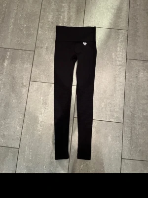 Svarta seamless leggings med ribbade detaljer - Svarta seamless leggings med hög midja och ribbade detaljer längs benen. Stretchigt material som sitter tight och bekvämt. Liten vit logga framtill vid midjan. Perfekta för träning eller chill.