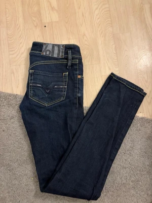 Mörkblå Diesel jeans W28 L34 - Säljer ett par mörkblå jeans från Diesel med klassisk femficksdesign och kontrastsömmar i gult. Modellen har raka ben och snygga detaljer vid fickorna, samt en liten Diesel-logga på myntfickan. Perfekta för dig som gillar stilrena jeans med bra passform.