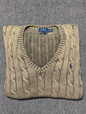 kabelstickad tröja Polo Ralph Lauren - Snygg beige kabelstickad tröja från Polo Ralph Lauren i mjuk pima bomull. Tröjan har v-ringad hals och långärmade ribbade muddar. Klassisk blå logga broderad på bröstet. Perfekt för dig som gillar stilrena och tidlösa plagg.