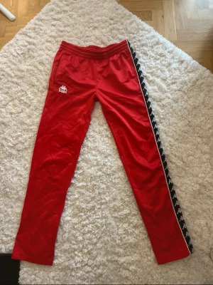 Röda Kappa byxor med knappar - Säljer ett par röda Kappa byxor i storlek small med svart och vit logoband längs sidorna. Byxorna har elastisk midja, fickor och tryckknappar längs ena benet för att kunna öppna upp. Perfekt för dig som gillar sportig och retro stil.