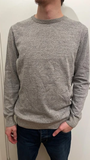 Grå stickad tröja med mockadetaljer - Snygg grå tröja från Springfield med rund hals och ribbade muddar. Tröjan har mörkbruna mockalappar på armbågarna som ger en cool touch. Perfekt för dig som gillar stilrena och bekväma plagg med lite extra detaljer.