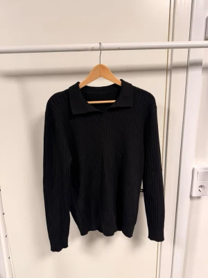 Svart ribbad pikétröja - Klassisk svart ribbad pikétröja med lång ärm och krage. Tröjan är stickad och har en clean look utan synliga knappar. Perfekt för dig som gillar minimalistisk stil och vill ha något som funkar året runt.
