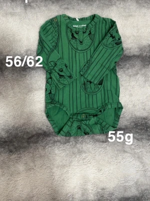 Grön body med djurmotiv från Mini Rodini - Säljer en grön body från Mini Rodini med svart tryck av djurmotiv och ränder. Bodyn har långa ärmar och praktiska tryckknappar i grenen. Perfekt för dig som gillar coola och unika prints på barnkläder.🌸Sparsamt använd. Storlek 56/62 