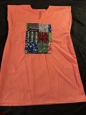 Korallfärgad afrikansk tunika med färgglad patch  - Säljer en korallfärgad blus med en unik fyrkantig patch framtill i olika mönster och färger som blått, grönt, svart, vitt och rött. Blusen har en enkel, rak passform och ett litet V-öppning vid halsen. Materialet känns mjukt och lätt, perfekt för dig som gillar färg och grafiska detaljer.