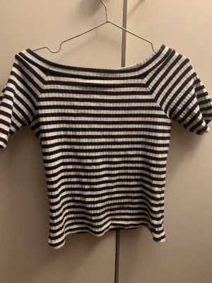 Randig ribbad topp från M&S Collection - Säljer en ribbad topp från M&S Collection med marinblå och vita horisontella ränder. Toppen har en bred, offshoulder-ringning och korta ärmar. Materialet är mjukt och stretchigt, perfekt för en avslappnad och trendig look. Storlek XS kanske passar till S