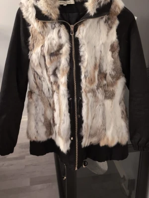 Pälsjacka från On Parle De Vous - Säljer en fluffig pälsjacka från On Parle De Vous i storlek 38. Jackan har svart tyg på ärmar och nederdel, samt vit, beige och grå fuskpäls framtill och på huvan. Stängs med dragkedja och har snörning i midjan för extra passform.