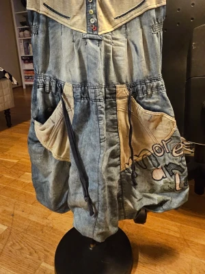 Desigual baggy jeansklänning med broderi - Unika baggy jeansklänning från Desigual i storlek 40. Klänningen har ljusblå denim med beige detaljer, stora fickor, snörning och broderad text på ena benet. Flera färgglada knappar framtill och cool patchwork-design ger en kreativ vibe.