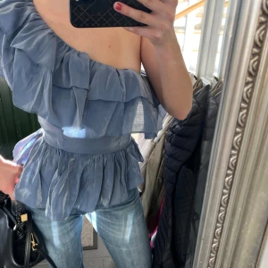 Blå oneshoulder blus med volanger - Säljer en snygg blå oneshoulder blus med volanger. Perfekt för en stilren look. Blusen har en midja som ger en fin passform och är tillverkad i ett lätt och luftigt material. Säljer då den var liten för mig i bysten (då jag har D-E kupa)