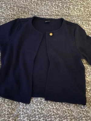 Mörkblå kofta med guldfärgad knapp - Snygg mörkblå kofta från Gina Tricot med rund halsringning och en guldfärgad knapp upptill. Koftan har korta ärmar och är tillverkad i mjukt material som känns skönt mot huden. Perfekt att slänga på sig när det är lite kyligt ute.