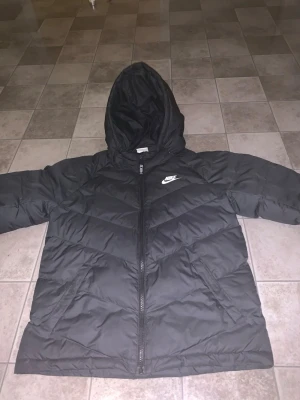 Svart pufferjacka från Nike, stl L - Svart pufferjacka från Nike med huva och dragkedja framtill. Jackan har ett stilrent, quiltat mönster och två sidofickor. Passar dig som är 147-158 cm. Perfekt för kalla dagar när du vill hålla dig varm och se sportig ut.