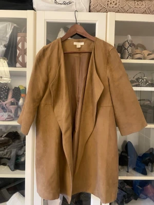Beige omlott kappa från H&M - Säljer en snygg beige kappa från H&M i storlek 38. Jackan har omlott-design med breda slag och trekvartsärmar. Materialet är mjukt och mockaliknande, vilket ger en chill och stilren vibe. Perfekt för dig som gillar minimalistisk look.