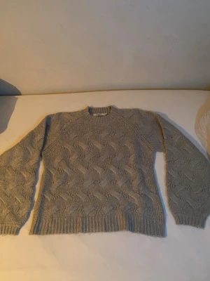 Grå stickad tröja från NA-KD - Säljer en grå stickad tröja från NA-KD i storlek S/M. Tröjan har ett snyggt kabelstickat mönster och ribbade muddar vid hals, ärmslut och nederkant. Perfekt för kyliga dagar och passar till jeans eller kjol.