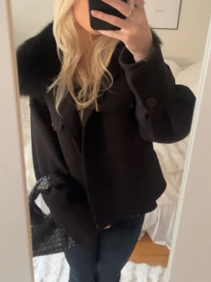 Svart kappa med pälskrage Meotine - Stilren svart kappa med bred pälskrage och stora knappar framtill. Kortare modell som ger en trendig siluett. Perfekt för dig som vill ha en klassisk men ändå modern look. Passar till både jeans och klänning.