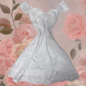 Vit offshoulder spetsklänning - Romantisk vit klänning med offshoulder-modell och stor rosett framtill. Klänningen har vackra spetsdetaljer och blommigt mönster över hela tyget. Perfekt för sommarens speciella tillfällen.