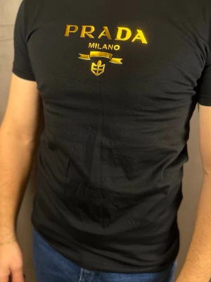 Svart t-shirt från Prada Milano - Snygg svart t-shirt från Prada Milano med guldfärgat tryck på bröstet. T-shirten har korta ärmar och en klassisk passform som sitter tight. Tillverkad i mjuk bomull som känns skön mot huden. Perfekt för dig som vill ha en stilren och exklusiv look.