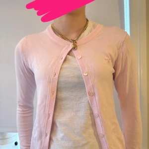 Ljusrosa cardigan från Gant - Söt ljusrosa cardigan från Gant med rund halsringning och knappar framtill. Liten broderad logga på bröstet och ribbade muddar vid ärmslut och nederkant. Perfekt att slänga över en t-shirt för en chill look.