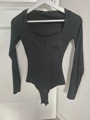 Svart ribbad body från ASOS DESIGN - Snygg svart body från ASOS DESIGN med lång ärm och markerade sömmar vid bysten. Modellen är ribbad och har en djup, fyrkantig ringning. Perfekt att styla med jeans eller kjol för en trendig look. Använd 1 ggr