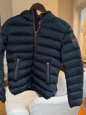 Svart pufferjacka från Colmar - Snygg svart pufferjacka från Colmar med huva och två dragkedjeförsedda fickor framtill. Jackan har en quiltad design med breda kanaler och en diskret logotyp på ärmen. Perfekt för kalla vinterdagar och riktigt skön att bära.