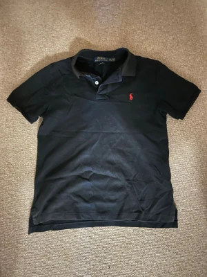 Svart pikétröja från Polo Ralph Lauren - Klassisk svart pikétröja från Polo Ralph Lauren med korta ärmar och krage. Tröjan har två knappar vid halsen och den ikoniska röda logotypen broderad på bröstet. Hör av dig vid minsta fundering!😊