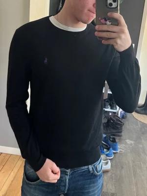 Svart tröja från Ralph Lauren - Klassisk svart tröja från Ralph Lauren med rund halsringning och diskret lila logga broderad på bröstet. Tröjan har långa ärmar och ribbade muddar vid ärmslut och nederkant. Perfekt till jeans för en clean och stilren look. Väldigt bra skick bara ett väldigt litet hål vid kragen syns på bilden.