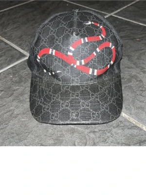 Gucci keps med orm-motiv - Snygg svart keps från Gucci med klassiskt GG-mönster och en röd orm broderad framtill. Kepsen har böjd skärm och justerbar rem bak. Materialet är canvas med exklusiv känsla. Perfekt för dig som vill sticka ut med en ikonisk accessoar. Äkta såklart 