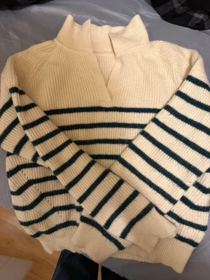 Randig stickad tröja med krage - Krämvit stickad tröja med mörkgröna horisontella ränder på ärmar och kropp. Tröjan har en hög krage med liten V-öppning framtill och ribbstickade muddar. Perfekt för en avslappnad och trendig look.