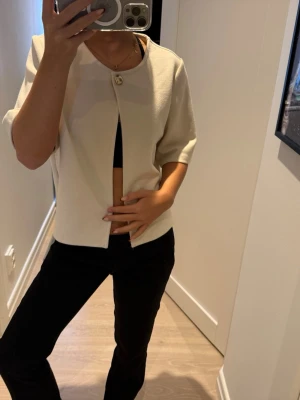 Krämvit kortärmad tröja från Gina tricot - Stilren krämvit blus från Gina Tricot i storlek M med en guldknapp som detalj