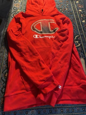 Röd hoodie från Champion - Snygg röd hoodie från Champion med stor logga på bröstet och klassisk huva. Mjuk bomullskänsla och känguruficka framtill. Perfekt för en avslappnad streetwear-look. Champion-logga även på ärmslutet.