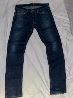Lee Mörkblå slim fit jeans med slitningar - Snygga mörkblå jeans i slim fit-modell med diskreta slitningar och klassiska fem fickor. Jeansen har kontrastsömmar och gylf med dragkedja och knapp. Perfekta för en trendig och avslappnad look.
