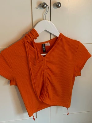 Orange ribbad croppad topp  - Trendig orange croppad topp från Gina Tricot med ribbad struktur. Toppen har korta ärmar, v-ringning och snörning framtill som ger en snygg rynkad effekt. Perfekt för dig som vill sticka ut med färg och stil. Snörena är tänkta att knyta runt magen. 