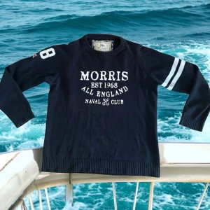 tröja från Morris - Mörkblå tröja från Morris med vit text och tryck på bröstet, samt siffran 68 och en liten brittisk flagga på ena ärmen. Tröjan har rund hals, ribbade muddar och två vita ränder på ena ärmen. Perfekt för en sportig och avslappnad stil.