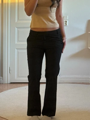 Svarta kostymbyxor med bootcut - Säljer ett par svarta kostymbyxor med bootcut-modell och låg midja. Byxorna har en klassisk look med raka ben som är lätt utsvängda nedtill. Materialet ser ut att vara en mjuk och följsam mix, perfekt för en clean och stilren outfit.